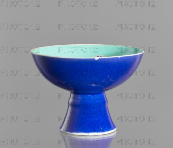 A porcelain footed bowl, Chinese, 19th century Qing Dynasty. Cobalt blue exterior glaze, green interior glaze; overglaze iron hieroglyphs, overglaze enamel paints, gilded edge with medal-gilding. Height 12 cm, diameter 16.9 cm., and provenance., A ceramic bowl on a pedestal with a glossy surface, showing a deep blue exterior and a light turquoise interior; the rim has a thin pale edge and a small chip visible, and the bowl sits on a flared base that narrows at the neck, all set against a neutral gray background with a subtle reflection beneath.