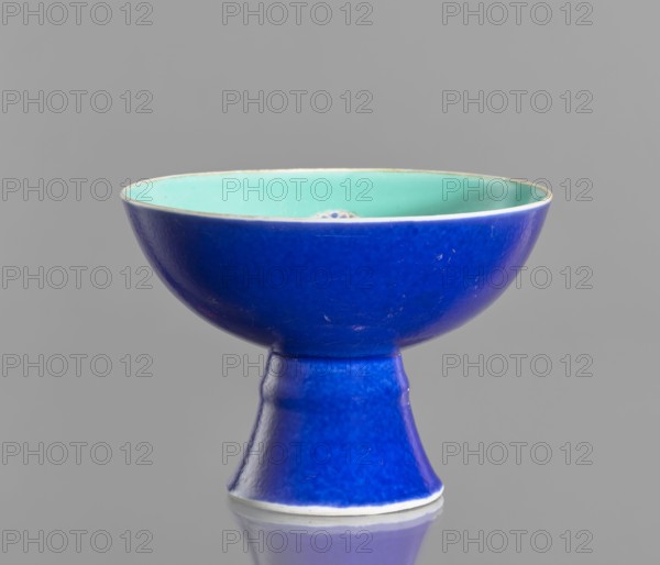 A porcelain footed bowl, Chinese, 19th century Qing Dynasty. Cobalt blue exterior glaze, green interior glaze; overglaze iron hieroglyphs, overglaze enamel paints, gilded edge and medal-gilding. Height 12 cm, diameter 16.9 cm., and provenance., A shallow bowl on a pedestal with a glossy blue exterior and a glossy mint green interior, sitting against a neutral gray background and reflected faintly on the surface below; the bowl shows some surface imperfections and subtle variations in the blue glaze.