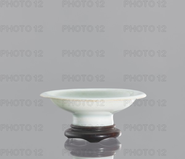 Alcohol bowl with base., Chinese; 19th century. Porcelain vodka bowl alcohol vessel with plate-like form: widely flared walls and a protruding rim, resting on a tall stem. Transparent bluish glaze. Flat round base with a side cut at the bottom into three flat legs; made of profiled wood. Height 3 cm; diameter 10.3 cm., A shallow, round ceramic dish with a wide flared rim sits on a dark wooden stand; the dish is a pale celadon green with subtle variations in tone and a slightly glossy finish, and the rim shows a faint warmer edge. The wooden stand is a deep brown with a smooth, polished surface and rounded feet. The objects are placed against a uniform light gray background that reflects faintly beneath the stand.