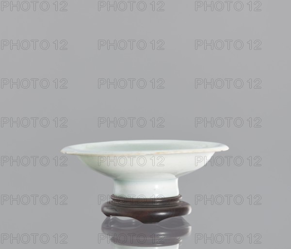 Alcohol bowl with base, Chinese, ceramics, porcelain vodka bowl. Plate with widely flared walls and a protruding rim. The bowl rests on a tall stem. Transparent glaze in a bluish tone. Flat, round base with a side cut at the bottom into three flat legs, made of profiled wood. 19th century, China. Vessel. Height 3 cm, diameter 10.3 cm., A shallow bowl with a wide rim and a short pedestal base sits on a carved stand; the bowl is a pale bluish-white color with subtle greenish tints and a thin slightly darker edge, while the stand is a dark brown wood; the background is a uniform light gray and the surface below the stand shows a soft reflection.