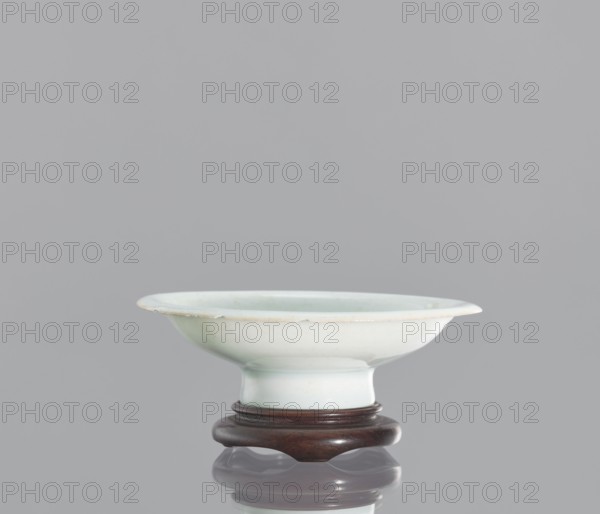 Alcohol bowl with base, Chinese, ceramics, porcelain vodka bowl. Plate with widely flared walls and a protruding rim. Bowl rests on a tall stem. Transparent bluish glaze. Flat, round base with a side cut at the bottom into three flat legs, made of profiled wood. 19th century, China. Vessel; height 3 cm, diameter 10.3 cm., A shallow ceramic bowl with a wide rim sits on a dark wooden stand; the bowl is pale celadon with subtle off-white and very light green tones and a smooth glossy finish, the rim shows a slightly warmer, faintly beige edge, and the wooden stand is a rich dark brown with a polished sheen, all set against a uniform light gray background.