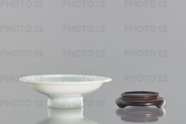Alcohol bowl with base, Chinese, 19th century. Porcelain vodka bowl with widely flared walls, a protruding rim, and resting on a tall stem. Transparent bluish glaze. Flat, round base with a side cut at the bottom into three flat legs; made of profiled wood. Height 3 cm, diameter 10.3 cm., and provenance., A shallow ceramic bowl with a pale greenish-white glaze sits on a raised foot beside a small round wooden stand with a dark brown finish; both objects cast soft reflections on the glossy surface beneath them, and the background is a uniform light gray.