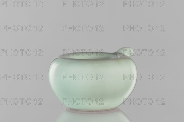 Water vessel, Chinese, Qing dynasty Kangxi period, 1662–1722. Ceramic celadon glazed decorative vessel, signed. Height 6.5 cm; width 11.7 cm., A smooth, glossy pale green ceramic vessel with a rounded, bulbous shape and a small, curved spout on one side, set against a neutral gray background and reflected faintly below; the surface shows subtle variations of lighter and slightly darker green and soft highlights.