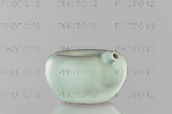 Water vessel, Chinese, Qing dynasty Kangxi period, 1662–1722. Ceramic celadon decorative vessel with celadon glaze. Signed. Height 6.5 cm, width 11.7 cm. Unknown provenance., A smooth, rounded ceramic vessel with a glossy pale green glaze sits against a neutral gray background, showing a subtle gradient of mint green to slightly darker green with faint warm brownish-green areas; the vessel has a small tubular spout on one side and a gently concave rim, and the surface reflects soft highlights and a faint shadow on the reflective surface beneath it.