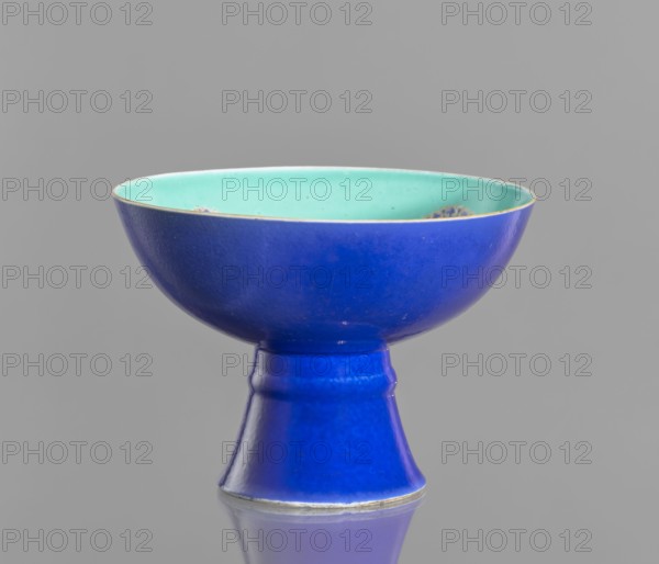 A tall footed porcelain bowl, Chinese, 19th century Qing Dynasty. Outer glaze stained cobalt, inner glaze green. Overglaze iron hieroglyphs, gilding, overglaze enamel paints, gilded edge. Height 11.8 cm, diameter 16.8 cm., and provenance., A shallow bowl with a flared foot, shown against a plain gray background, featuring a deep cobalt blue exterior and foot and a pale turquoise interior; the rim has a thin metallic edge and there are a few small blemishes and speckles on the interior surface, while the reflective surface beneath the object shows a muted blue reflection.