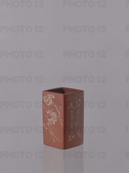 Vessel, Japanese art, Japanese culture, engraved decorations, ceramics, flowers iconography, red-brown stoneware; engraved decoration dyed white with engobe, 19th century, Japan, vessel; height 11.1 cm, width 6.3 cm, depth 6.3 cm, A rectangular reddish-brown ceramic vessel with an open top sits on a reflective surface, showing a faint mirrored outline beneath it; one visible side features an engraved floral motif and another side shows an engraved vertical pattern; the background is a smooth pale grayish-lavender color and the vessel's interior appears darker brown.