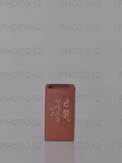 Vessel, Japanese art, Japanese culture, engraved decorations, ceramics, flowers iconography, unknown provenance; red-brown stoneware with engraved decoration dyed white with engobe; 19th century, Japan; vessel; height 11.1 cm, width 6.3 cm, depth 6.3 cm, A rectangular reddish-brown ceramic vessel stands upright on a reflective surface against a uniform light gray background, showing a vertical face with a few pale marks of decoration; the top is open revealing a darker interior, and the piece casts a subtle reflection and shadow on the surface.