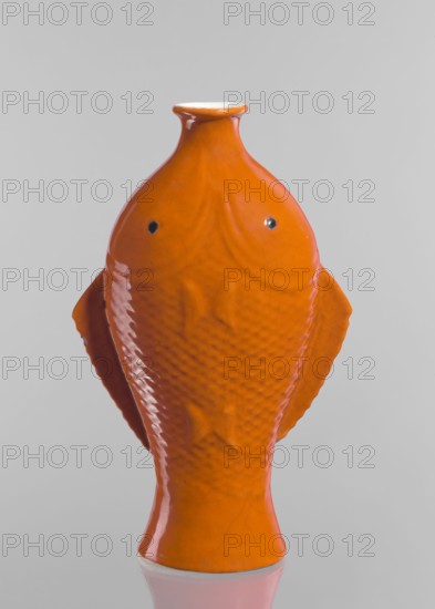 Vase in the shape of two fish, Chinese, Qing dynasty 1644–1911, ceramics decorative vessel with red and white glaze, 19th century, China; vessel, height 35.8 cm, An orange ceramic object shaped like a stylized fish stands upright against a pale gray background, showing a glossy surface with textured scale-like patterns, a narrow neck and flared rim at the top, two small dark circular details near the upper front, and paired flattened fins extending from the sides, with reflections visible on the smooth finish.