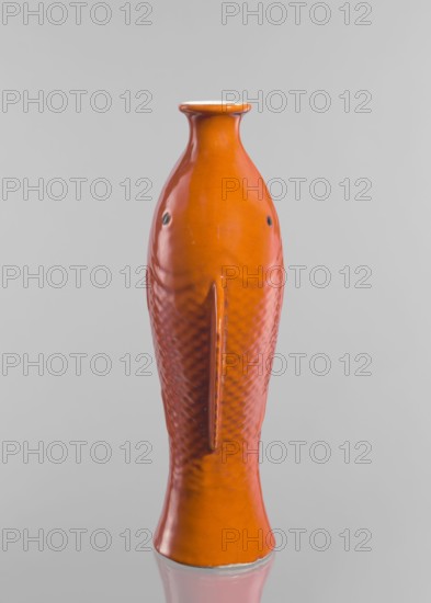 Vase in the shape of two fish, Chinese, Qing dynasty 1644–1911, ceramics, decorative vessel with red and white glaze, 19th century, China; height 35.8 cm, An orange ceramic object shaped like a fish with a narrow neck and flared rim, showing a textured scale pattern along its body, small dark circular details where eyes would be, a raised vertical ridge along the center of the body, and a glossy surface that reflects light against a light gray background with a faint reflection beneath it.