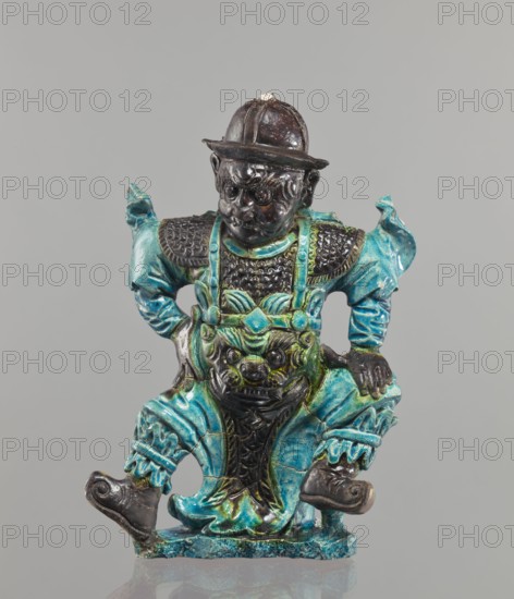 Warrior figurine, Chinese, ceramic sculpture/figurine of a male warrior, green and brown glaze, 17th century, height 30.5 cm. Creator, provenance unknown., A glazed ceramic figurine of a seated warrior-like figure wearing a rounded hat and ornate armor, with a prominent stylized face on the torso. The figure shows a glossy dark brown head and hands, and vivid turquoise and teal garments and decorative elements, with areas of greenish glaze and darker brown or black accents on the armor, facial features, and footwear. The posture has the figure’s legs bent and feet turned outward on a turquoise-toned base, with textured detailing on the shoulders, chest, and pants, and a carved expressive face on the central apron area. The background is a smooth pale gray.