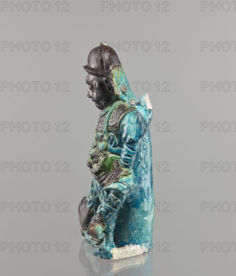 Warrior figurine, Chinese, ceramic figurine of a male warrior, green and brown glaze, 17th century, China; sculpture, height 30.5 cm, A glazed ceramic figurine showing a standing figure in profile against a plain gray background, with a dark brown head and helmet, a turquoise and teal robe with green accents, and areas of darker brown on the hands and lower legs; the figure wears layered armor pieces and a hood or cape, and the surface shows glossy, uneven glaze with some worn or chipped spots revealing lighter material at the base.
