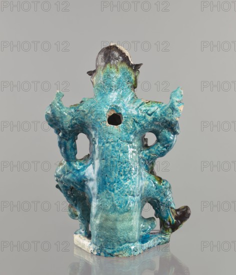Warrior figurine; creator, Chinese; ceramic figurine of a male warrior with green-brown glaze; 17th century ; China; sculpture; height 30.5 cm., The image shows a sculptural object photographed against a neutral gray background, featuring a glossy, irregular form with a predominantly turquoise and teal glaze that has areas of lighter aqua and hints of darker blue; there are also small patches of brown, black, and green on the upper and lower parts, and some exposed beige or off-white areas where the glaze is missing. The object has a vertical back view with asymmetrical protrusions and rounded, organic shapes, including a central hollow opening and several rounded perforations; the surface shows a crackled texture and uneven glazing with drips and pooling, and the piece sits on a small square base with a reflective surface below it that creates a subtle reflection.