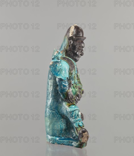 Warrior figurine, Chinese; ceramic sculpture of a male warrior with green-brown glaze; 17th century, height 30.5 cm; maker, provenance unknown., A glazed ceramic figurine shown in profile with a turquoise and blue robe, green and teal accents, and a dark brown face and hands; the figure wears a dark brown rounded hat with a lighter patch on top and has sculpted facial features and curly hair; the robe has visible folds and a cape-like back, with areas of lighter blue and mottled texture; the hands rest near the front of the body holding a textured object with green and brown tones; the base shows worn edges with exposed ceramic in beige and brown; the background is neutral gray.