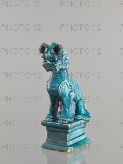 Lion fo; maker, creator role/craftsman, provenance unknown. Chinese, Qing dynasty 1644–1911. Ceramic animal figurine lion/fo, sea-green glaze. Sculpture/figurine, height 28 cm; 17th century ., A glossy turquoise ceramic figurine of a seated mythical creature with a curled mane, prominent ears, and stylized facial features sits on a stepped rectangular base; the surface shows areas of wear with exposed darker and brownish patches, and the scene includes shades of turquoise, blue, brown, and gray against a plain light gray background.