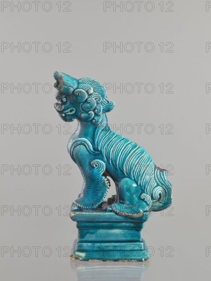 Lion fo, creator unknown, Chinese, Qing 1644–1911, 17th century, ceramic animal figurine of a lion/fo with sea-green glaze, sculpture/figurine, height 28 cm., A turquoise ceramic figurine of a stylized lion-like creature sits on a rectangular base against a plain light gray background, the creature shown in profile facing left with a curled mane made of carved swirls and ridged fur along its back and tail, rounded paws with visible toe details, a prominent snout and open mouth, and areas of minor wear revealing a darker underlying material at edges; colors visible include turquoise, darker teal accents in recesses, small areas of brownish wear, and the light gray background.