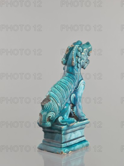 Lion fo, Chinese, Qing 1644–1911, 17th century, ceramic sculpture/figurine of a lion fo, sea green glaze, height 28 cm, animal figurine, iconography: fo., A turquoise-blue ceramic figurine of a seated mythical creature on a square pedestal, shown from the rear and slightly in profile; the creature has a mane of stylized curls and ridged, flowing fur along its back and tail, with visible worn areas where the glaze is chipped to reveal a darker underlying material; the pedestal and base share the same turquoise-blue color with lighter and darker variations, and the scene sits against a pale gray background with a subtle reflection on the surface beneath the pedestal.