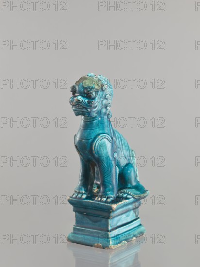 Lion fo, Chinese, Qing 1644–1911, 17th century, ceramics sculpture figurine of a lion fo, sea green glaze, height 27 cm, animal figurine, China, ceramics, iconography: fo lion, A small sculpted figure of a seated mythical lion-like creature with a stylized mane and curled details, shown on a stepped rectangular base; the figure is glossy and predominantly turquoise blue with areas of darker teal and traces of worn lighter spots, and there are patches of a greenish patina on the head and small exposed areas of a brownish or tan underlying material at edges and corners; the creature has pronounced facial features, a ridged back, and detailed paws resting on the base, all set against a plain light gray background.