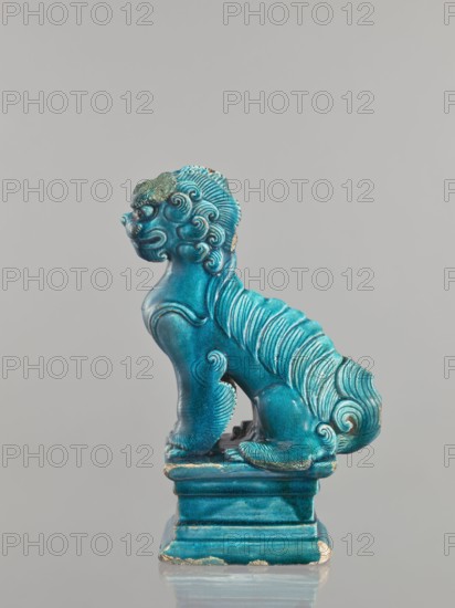 Lion fo, Chinese, Qing 1644–1911, 17th century, ceramic animal figurine of a lion fo with sea green glaze, sculpture, height 27 cm, A glazed ceramic statue of a mythical lion-like creature in a seated pose on a rectangular plinth, shown in profile facing left, with a stylized mane of carved waves and curls, a rounded snout, pronounced brow, curled tail, and detailed paws. The surface is a vibrant turquoise-blue glaze with areas of darker blue shading and small patches where the glaze is worn, revealing a pale underlying material. The background is a uniform light gray.