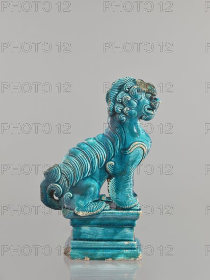 Lion fo, creator unknown, Chinese, Qing dynasty 1644–1911, 17th century, ceramic animal figurine of a fo lion with sea-green glaze, sculpture, height 27 cm., A small sculpted figure of a stylized animal with a curled mane and tail sits on a rectangular base, rendered in a bright turquoise-blue glaze with patches of darker blue-green and small areas where the glaze is worn to reveal a pale underlying material; the animal displays flowing carved ridges along its body and limbs, a pronounced snout and open mouth, rounded ears, and decorative spiral motifs in its mane and fur, while the base shows stepped molding and slight chipping at the edges; the background is a uniform light gray.