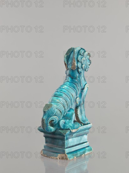 Lion fo, Chinese, Qing 1644–1911, 17th century, ceramic animal figurine lion/fo with sea green glaze, sculpture, height 27 cm., A sculptural figure of an animal-like creature seated on a stepped base, shown from behind and slightly to one side, with carved, flowing ridges and curled decorative fur details along its back, tail, and head; the piece is predominantly turquoise blue with areas of darker teal and patches where the surface is worn to a rough, beige-gray material; the base shares the same turquoise-blue glazing with some worn edges revealing the underlying beige-gray, and the object sits against a plain light gray background.