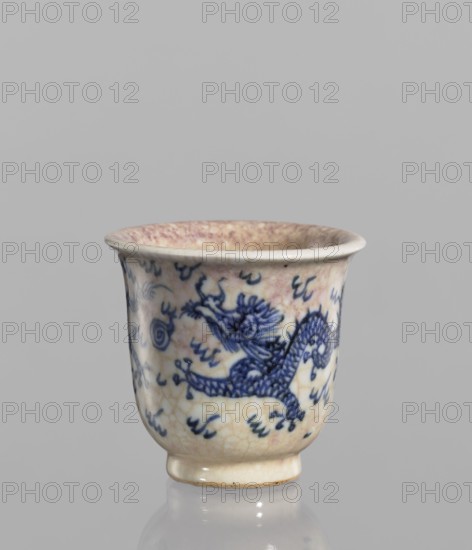 Bowl, Chinese, blue-and-white porcelain, 19th century. Bowl with a rolled edge and a small foot; crackled glaze stained with pink and lilac spots; decoration includes dragons, flames, cracklework; pearls motif. Vessel, height 7.4 cm, diameter 7.8 cm., A small ceramic cup with a flared rim and a short foot, predominantly off-white with a crackled glaze showing faint beige and pinkish tones inside the rim, decorated on the exterior with a blue painted dragon motif and scattered blue brushstroke marks; the surface shows age-related crazing and slight discoloration, and the cup sits on a reflective surface against a plain light gray background.