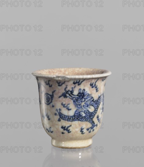Bowl, Chinese, blue-and-white porcelain, 19th century. Bowl with a rolled edge and a small foot; crackled glaze stained with pink and lilac spots; decoration includes dragons, cracklework, and flames. Vessel, height 7.4 cm, diameter 7.8 cm., A small ceramic cup with a flared rim and a short foot, showing a crackled cream-colored glaze with areas of beige and light brown toning, decorated with blue painted motifs including a stylized dragon and flowing cloud-like marks; the rim has a small chipped area with exposed ceramic, and the cup sits on a reflective gray surface against a plain gray background.