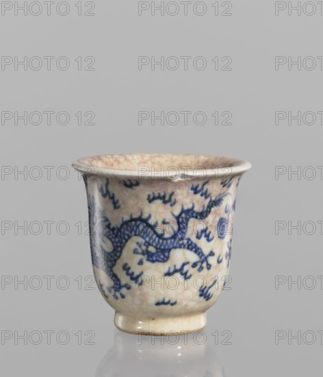 Bowl, Chinese, blue-and-white porcelain with crackled glaze stained with pink and lilac spots; decorated with dragons and flames; rolled edge and small foot; 19th century; height 7.4 cm, diameter 7.8 cm., A small ceramic cup with a flared rim and short foot, showing a crackled cream-colored glaze with areas of light beige and pale pink toning; the surface is decorated with a group of blue brush-painted motifs including a sinuous dragon-like form and scattered short strokes; the rim has slight wear and a small chip, and the cup sits on a reflective surface that produces a soft mirrored shape; visible colors include cream, beige, pale pink, blue, and a hint of gray in the background.