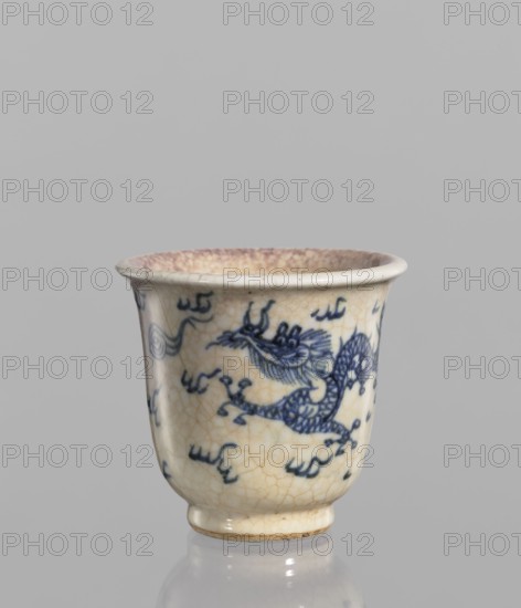Bowl, Chinese, blue-and-white porcelain bowl with a rolled edge and small foot. Crackled glaze stained with pink and lilac spots, decorated with pearls, dragons, cracklework and flame motifs. 19th century, China. Height 7.4 cm; diameter 7.9 cm., A small ceramic cup sits against a neutral gray background, showing a rounded flared rim and a slightly tapered body that narrows toward a short foot. The cup's surface is a creamy off-white with a network of fine crackle lines and areas of subtle age-toned discoloration. A decorative motif in deep blue wraps around the exterior, forming sinuous, scaly shapes resembling a dragon with flowing whiskers and a curling tail, interspersed with short curved brushstrokes. The rim interior shows a faint wash of muted purplish-beige, and the foot edge has a slightly darker beige band. Reflections on the glossy glaze create small highlights on the cup's body and base.