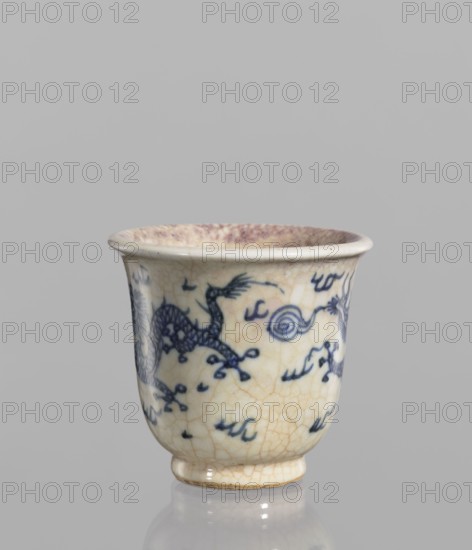 Bowl, Chinese, 19th century. Blue-and-white porcelain bowl with rolled edge and small foot; crackled glaze stained with pink and lilac spots. Decoration: pearls, dragons, flames; cracklework. Vessel; height 7.4 cm, diameter 7.9 cm. Unknown provenance., A small ceramic cup with a flared rim and a short foot, showing a crackled cream glaze with blue painted decorative motifs that include a dragon-like creature and swirling cloudlike shapes; the interior rim has a faint purplish-brown discoloration, and the cup sits on a reflective neutral-gray surface against a plain light-gray background.