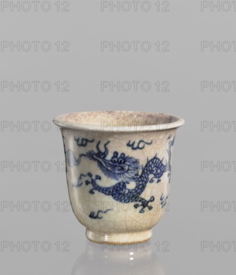 Bowl, Chinese, blue-and-white porcelain, crackleware decoration with flames, dragons and pearls iconography; rolled edge and small foot; crackled glaze stained with pink and lilac spots; 19th century, China; vessel; height 7.4 cm, diameter 7.9 cm., A small ceramic cup with a slightly flared rim and a short foot, showing a crackled off-white glaze with visible crazing and areas of light beige and tan discoloration; the cup is decorated with a flowing blue painted dragon motif and additional small blue cloud-like brushmarks around it, and it sits on a reflective gray surface against a plain light gray background.