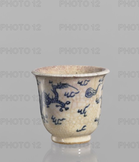 Bowl, Chinese, ceramic; blue-and-white porcelain with cracklework glaze stained with pink and lilac spots; decorated with pearls, dragons, and flames; rolled edge and small foot; 19th century, China; vessel; height 7.4 cm, diameter 7.9 cm., A small ceramic cup with a flared rim and a short rounded foot, showing a crackled glaze and an aged surface. The cup's body is mainly creamy off-white with areas of warm beige and pale yellowing, and it bears hand-painted blue decorative marks and motifs across its exterior. The interior shows a subtle pinkish-beige tone and visible crackling in the glaze. The overall scene has a neutral gray background and the cup sits on a reflective surface that echoes its colors.