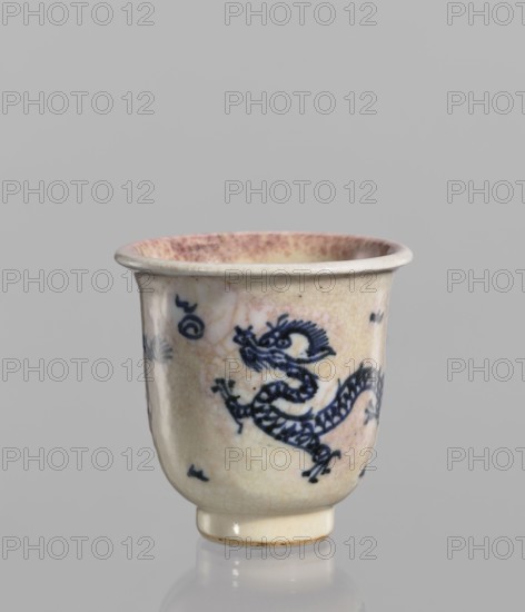 Bowl, Chinese, 19th century. Blue-and-white porcelain with cracklework decoration depicting dragons, pearls, and flames. Rolled edge and small foot; crackled glaze stained with pink and lilac spots. Vessel; height 7.7 cm, diameter 7.9 cm., and provenance., A small ceramic cup with a rounded body and a flared rim sits against a plain gray background, the cup’s surface showing a crackled glaze in off-white and pale beige with areas of pinkish staining inside the rim and near the upper exterior; a dark blue stylized dragon motif wraps around the body with additional small decorative marks in the same dark blue, and the cup rests on a short cylindrical foot with a slightly darker base edge.
