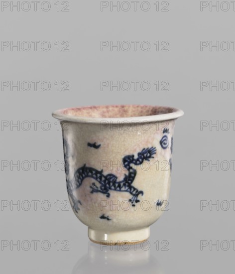 Bowl, Chinese, blue-and-white porcelain with cracklework decoration featuring dragons, pearls, and flames. Bowl with a rolled edge and a small foot; crackled glaze stained with pink and lilac spots. 19th century, China. Height 7.7 cm, diameter 7.9 cm., A small ceramic cup sits upright on a reflective surface against a plain light gray background, showing a curved rim and a short foot. The cup's outer surface is a crackled off-white with areas of pale pinkish wash, and it is decorated with a hand-painted dark blue serpentine dragon motif and a few small abstract marks. The interior rim shows a mottled pink and cream pattern. The foot has a slightly unglazed beige edge.