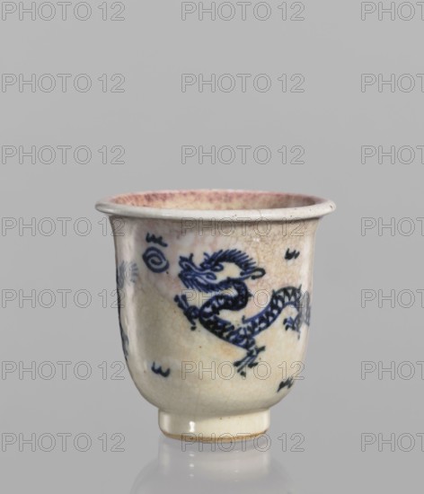 Bowl, Chinese, 19th century. Blue-and-white porcelain bowl with rolled edge and small foot. Crackled glaze stained with pink and lilac spots. Decoration: dragons, pearls, and flames. Vessel; height 7.7 cm, diameter 7.9 cm., A small ceramic cup with a flared rim and a short foot, showing a crackled cream-colored glaze with areas of pinkish discoloration near the rim, decorated with a dark blue stylized dragon motif wrapping around the body and a few small dark blue decorative marks, photographed against a plain light gray background.