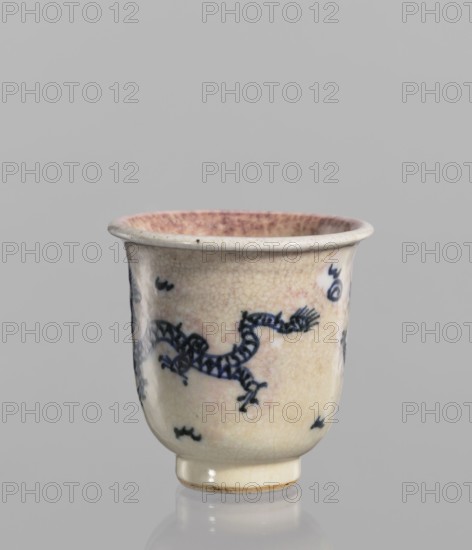 Bowl, Chinese, 19th century. Blue-and-white porcelain with cracklework decoration depicting dragons, pearls, and flames. Rolled edge and small foot; crackled glaze stained with pink and lilac spots. Vessel; height 7.7 cm, diameter 7.9 cm., and provenance., A small ceramic cup with a rounded body and short foot, shown against a plain light gray background, has a crackled off-white glaze with a warm beige and faint pinkish tint, and a few areas of darker beige spotting; a hand-painted dark blue sinuous dragon motif wraps around the body with several small dark blue decorative marks scattered nearby; the rim interior shows a denser pinkish wash, and the foot rim has a slightly darker beige edge.