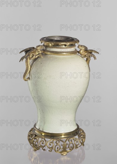 Vase, Chinese, Qing dynasty 1644–1911. Gilded bronze on the base and neck. Shoulders with two gilded bronze winged dragon figurines and a snake figurine. Underglaze engraved belly decoration with transparent greenish glaze showing plant-floral tendrils, dragons, snakes, and plant ornaments. Decorative vessel, 19th century, China; height 45.7 cm., A pale green ceramic vase with subtle embossed swirling patterns sits on an ornate gold-colored metal stand; the vase has a rounded body tapering toward the base and a short neck topped with a metal rim, and there are decorative gold-colored metal mounts at the shoulder shaped like stylized animal heads with wing-like extensions.