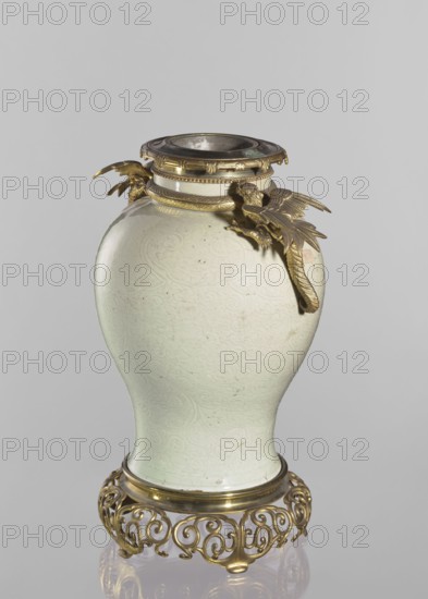 Vase, Chinese, Qing dynasty 1644–1911. Gilded bronze on the base and neck. Shoulders with two gilded-bronze winged-dragon figurines and a snake figurine. Underglaze engraved belly decoration with transparent greenish glaze showing plant-floral tendrils. Decorative vessel featuring dragons, snakes, and plant ornaments. 19th century, China. Height 45.7 cm., A pale greenish-white ceramic vase with a bulbous body and narrow neck sits on an ornate golden metal stand; the vase surface shows subtle raised swirling patterns and a few small dark specks. The neck of the vase is fitted with a decorative golden metal collar and a removable rim piece with a darker inner ring, and two sculpted golden metal bird-like figures with spread wings are attached near the neck, their textured feathers and curved beaks extending outward. The golden stand beneath the vase features an openwork scroll motif and rounded feet, and the overall color palette includes pale greenish-white, gold, and hints of darker me...