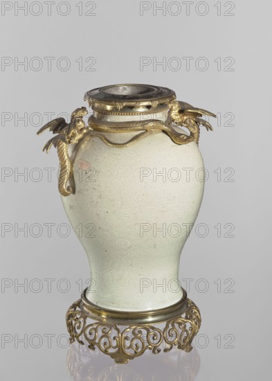 Vase, Chinese, Qing dynasty 1644–1911. Decorative vessel with dragon and snake iconography and plant/floral ornaments. Gilded bronze on the base and neck. Shoulders with two gilded bronze winged dragon figurines and a snake figurine. Underglaze engraved belly decoration with transparent greenish glaze showing plant-floral tendrils. 19th century, China. Vessel, height 45.7 cm., A pale celadon vase with a subtle swirling embossed pattern sits on an ornate gilded metal stand; the vase has a rounded body that tapers toward the base and a wide neck fitted with a decorative metal collar, and a pair of gilded dragon-shaped handles with textured wings and sinuous bodies wrap around the upper portion of the vase, showing metallic gold highlights against the pale green-white ceramic, with additional gold tones on the collar and stand, and a neutral gray background.