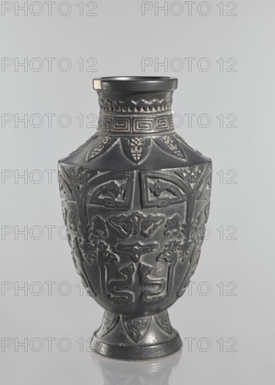 Vase, Japanese, Meiji 1868–1912–Taisho 1912–1926, first half of the 20th century. Ceramic vessel from Kagoshima, height 42 cm. Vase on a slightly widening foot with clearly defined shoulders and a short neck; dark brown glaze; decorated with meander, plant and animal ornaments; signed., A single dark gray vase with a high neck and flared rim stands against a light gray background, showing an overall matte surface with slightly lighter worn areas; the body is decorated with raised ornate patterns including geometric bands near the neck, stylized leaf or petal shapes at the shoulder, and a variety of abstract and symmetrical relief motifs along the midsection and lower body; the base flares outward and features repeating decorative elements that echo the shoulder motifs; the vase casts a faint reflection below it on the surface it stands on.
