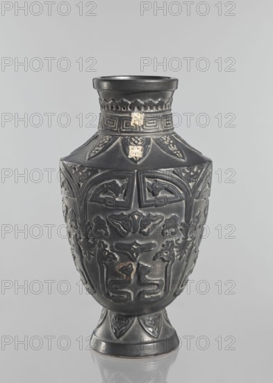 Vase, Japanese, Meiji–Taisho period late 19th–early 20th century, vessel; slightly widening foot, clearly defined shoulders and short neck; dark brown glaze; decorated with meander, plant and animal motifs; signed; 1st half of the 20th century; Kagoshima, Japan; height 42 cm., A dark gray ornamental vase with a narrow footed base widening into a bulbous body and tapering to a short neck and flared rim, decorated with raised, symmetrical carved motifs and stylized animal and abstract patterns, with a few small worn or chipped areas showing lighter, almost beige patches, set against a light gray background.