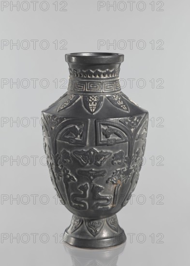 Vase, Japanese, Meiji–Taisho period late 19th–early 20th century. Ceramic decorative vessel from Kagoshima, Japan. Vase on a slightly widening foot with clearly defined shoulders and a short neck; dark brown glaze; signed. Height 42 cm. Subjects: meander ornament, plant ornament, animals., A dark-colored decorative vase stands against a light gray background; the vase has a narrow neck flaring to a wider rim and a rounded body that tapers to a short pedestal base, all covered with raised ornamental patterns including repeating geometric bands, stylized motifs, and curving relief designs; the surface appears smooth and slightly reflective with some lighter wear spots, and the visible colors are dark gray to black with small areas of lighter gray and hints of brownish wear.