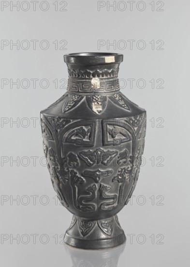 Vase, Japanese, Meiji–Taisho periods late 19th–early 20th century; 1st half of the 20th century. Ceramic decorative vessel from Kagoshima, Japan. Vase on a slightly widening foot with clearly defined shoulders and a short neck; dark brown glaze; decorated with meander, plant and animal motifs; signed. Height: 42 cm., A dark vase with a matte black surface sits against a light gray background, showing a rounded body that tapers to a narrow neck and flared rim and rests on a short pedestal base; the vase is decorated with raised, carved motifs forming symmetrical panels of stylized animal and abstract designs, with ornamental borders near the neck and base. Several small areas of lighter wear or residue appear on the neck and upper body, showing a pale beige contrast against the black. The overall visible colors are black, light gray background, and pale beige in a few worn spots.