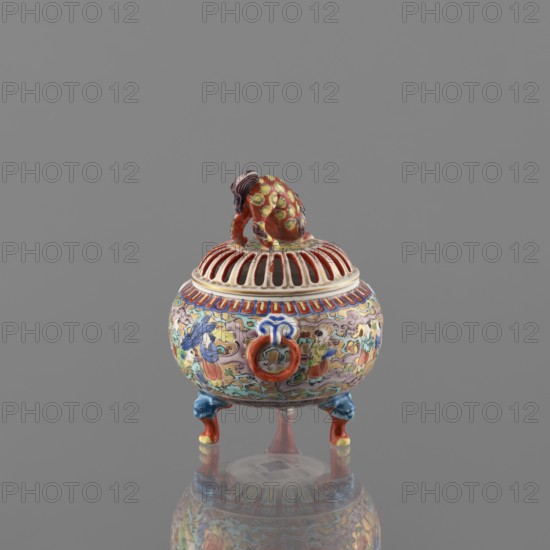 Incense burner with lid, Kutani, figurative scenes, Japanese art, Japanese culture, three-legged vessel, colored porcelain, Meiji 1868–1912, deer, porcelain pottery, grayish white glaze with a bluish tint, overglaze decoration in green, purple, red, blue, yellow with black outlines, 19th/20th century Meiji period, Japan, Kaga province, vessel, incense burner, height 12 cm, diameter 9 cm, A small decorative vessel sits on a reflective surface against a plain gray background, with a domed lid featuring open slats and a small sculpted animal figure perched on top. The vessel has a rounded body on short ornate legs and ring handles on its sides. Its surface is richly painted with intricate scenes and decorative motifs in colors including red, blue, yellow, green, pink, and white, with accents of darker tones. The sculpted animal on the lid is painted predominantly red with yellow and green patterning and darker hair details, and the legs show blue and red coloring with touches of yellow. T...