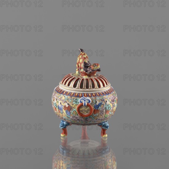 Incense burner with lid, Kutani maker/manufacturer, figurative scenes, Japanese art, Japanese culture, three-legged vessel, colored porcelain, Meiji 1868–1912, deer motif, porcelain pottery, grayish white glaze with a bluish tint; overglaze decoration in green, purple, red, blue, yellow with black outlines; 19th/20th century Meiji period; Japan, Kaga; vessel incense burner; height 12 cm, diameter 9 cm, A small ornate ceramic vessel with a domed, slotted lid topped by a sculpted animal figure sits on a reflective surface against a plain gray background; the piece is brightly painted with colors including red, blue, yellow, green, orange, pink, white, and black, featuring a band of vertical red and white stripes on the lid, a decorative rim with red and blue accents, figural and cloudlike painted scenes around the body in pastel and saturated hues, a prominent circular applied ring motif in red and blue on the front, and three short legs painted blue with red and yellow highlights, while...