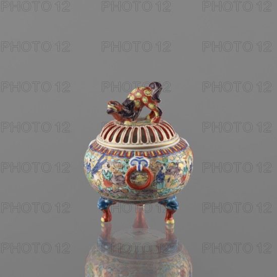 Incense burner with lid, Kutani, figurative scenes, karashishi iconography, deer; colored porcelain, Meiji 1868–1912, 19th/20th century; Kaga, Japan. Porcelain body, grayish, white glaze with a bluish tint. Decoration painted with overglaze paints in green, purple, red, blue, yellow with black outlines. Vessel incense burner, height 12 cm, diameter 9 cm., A small ornate ceramic vessel with a domed, slotted lid topped by a sculpted animal figurine, standing on three short curved legs; the vessel displays a complex painted surface with a group of human and animal figures and decorative motifs in a palette that includes red, blue, yellow, green, purple, orange, white, and black, and the lid features red and white ribbed openings beneath the animal, which is painted with yellow spots on a red body and dark purple mane; the legs are painted blue and red with yellow accents and the overall object rests on a reflective gray surface with a plain gray background.