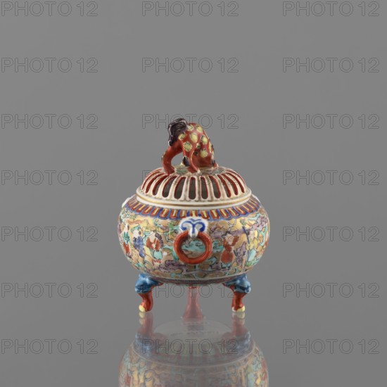 Incense burner with lid, Kutani, figurative scenes, Japanese art, Japanese culture, colored porcelain, karashishi iconography, deer, Meiji 1868–1912, porcelain body, grayish, white glaze with a bluish tint; decoration painted with overglaze paints: green, purple, red, blue, yellow in black outlines; 19th/20th century Meiji period; Japan, Kaga; vessel, incense burner; height 12 cm, diameter 9 cm., A small ornate round vessel with a domed pierced lid topped by a crouching animal figure, the object decorated all over with intricate painted motifs in red, blue, yellow, green, and creamy white; the lid has ribbed openings and the animal finial shows red and dark brown with yellow spots, while the body displays a banded rim in red and blue and multicolored scene-like patterns; the vessel stands on a group of small curved legs painted in blue and red with touches of yellow, and it sits on a reflective surface against a plain gray background.