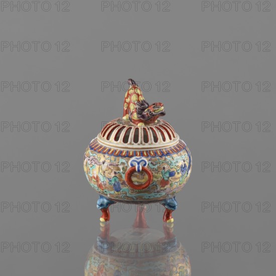 Incense burner with lid, Kutani, figurative scenes, Japanese art, Japanese culture, colored porcelain, karashishi iconography, deer, Meiji 1868–1912, porcelain body, grayish, white glaze with a bluish tint; decoration painted with overglaze paints: green, purple, red, blue, yellow in black outlines; 19th/20th century Meiji period, Japan, Kaga province; vessel, incense burner; height 12 cm, diameter 9 cm, A small ornate ceramic vessel sits against a plain gray background, featuring a rounded body on three short tripod legs with curled ends, each leg painted in blue and red with yellow highlights. The body is decorated all around with a busy scene rendered in multiple colors including blue, green, yellow, red, pink, white, and black, and it has a raised circular ring ornament on the front painted red with blue accents. The domed lid has vertical open slits and a banded rim with red and white details, topped by a sculpted mythical creature in red and yellow with black and purple painted f...