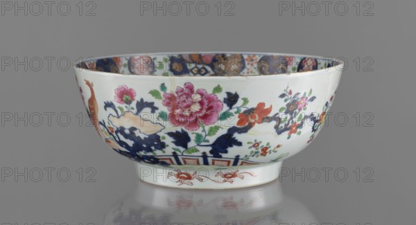 Bowl, Chinese, 19th century. Colored porcelain with grey-white glaze. Bowl on a flat-rimmed foot. Decoration: inside, a projection of peony sprigs at the bottom and a border with a pomegranate below the rim; outside, a loose composition of a garden scene with a fence, a peony bush, rocks, and two phoenixes fenghuang. Floral decorations, peonies; phoenixes iconography. Vessel; height 13.5 cm, diameter 32 cm., A porcelain bowl with a rounded shape and a short foot, decorated with painted floral motifs and foliage in colors including white, pink, red, green, dark blue, orange, and black, showing a large pink bloom and several smaller flowers and leaves around it, with dark blue cloud-like or rock-shaped forms near the base and a stylized bird figure at one side; the interior rim displays a patterned band in assorted colors, and the bowl sits on a reflective gray surface against a plain gray background.