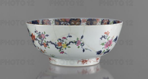 Bowl, Chinese, 19th century. Colored porcelain with grey-white glaze. Bowl on a flat-rimmed foot. Decoration: inside, a projection of peony sprigs at the bottom and a border with a pomegranate below the rim; outside, a loose composition of a garden scene with a fence, a peony bush, rocks, and two phoenixes fenghuang. Floral decorations; peonies, phoenixes iconography. Vessel, height 13.5 cm, diameter 32 cm., A shallow ceramic bowl with a white background sits against a gray backdrop, decorated with a floral motif that includes a few pink, red, yellow, green, and dark blue elements, and a band of multicolored patterned decoration visible along the inner rim; the bowl shows visible repair lines and a reflection beneath it on the surface.