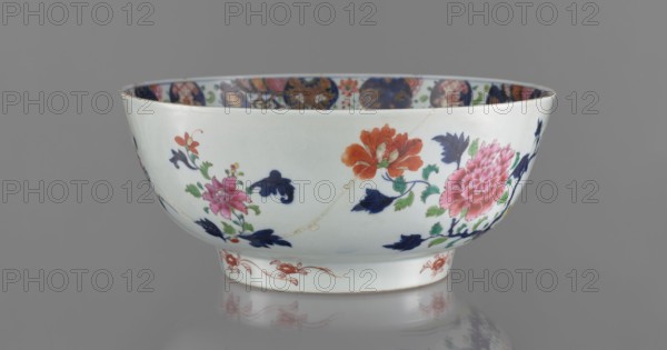 Bowl, Chinese, 19th century. Colored porcelain with grey-white glaze. Bowl on a flat-rimmed foot; height 13.5 cm, diameter 32 cm. Decoration: inside — projection of peony sprigs at the bottom and a border with a pomegranate below the rim; outside — loose garden scene with a fence, peony bush, rocks, and two phoenixes fenghuang. Floral decorations, peonies; phoenixes iconography., Function: vessel., A shallow porcelain bowl with a glossy white surface sits against a neutral gray background, decorated with a floral pattern featuring a few large blossoms in pink and orange, several dark blue stylized leaves, and green foliage; the interior rim shows a band of multicolored round motifs with orange, blue, green, and pink tones, and the foot of the bowl has a delicate red decorative band.